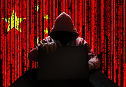 A hooded figure sitting at a desk with a laptop, against a red background featuring the Chinese flag